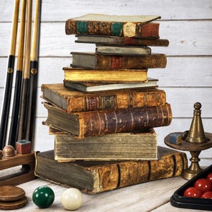 May include: A tall stack of weathered, antique hardcover books with worn leather spines and gold-gilded edges. Beside the books are three wooden pool cues, a small brass pedestal, a green billiard ball, a white cue ball, and red billiard balls.