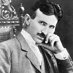 May include: A black and white portrait of a person with dark hair and a mustache, wearing a textured blazer over a white collared shirt. The person is positioned with one hand resting near their chin, looking directly toward the camera lens.