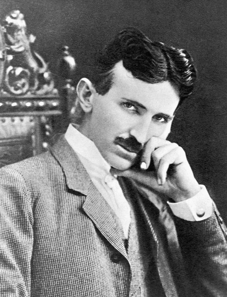 May include: A black and white portrait of a person with dark hair and a mustache, wearing a textured blazer over a white collared shirt. The person is positioned with one hand resting near their chin, looking directly toward the camera lens.