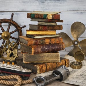 May include: A stack of antique books sits centrally, surrounded by a wooden ship wheel, a brass sextant, a model ocean liner, a rope, a metal piston, a brass boat propeller, and a compass on a nautical map against a white wooden background.