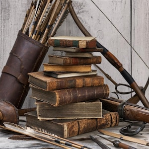May include: A stack of weathered, antique hardback books with worn leather spines and aged pages. Beside the books sits a brown leather quiver filled with feathered arrows and a wooden recurve bow with a dark grip, arranged in a rustic composition.