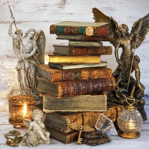 May include: A stack of antique books sits between two angel statues, one light gray and one bronze. A small cherub figurine, a gold-toned candle holder, a glass lantern, a gold key with wings, and a clear crystal prism are arranged in front.