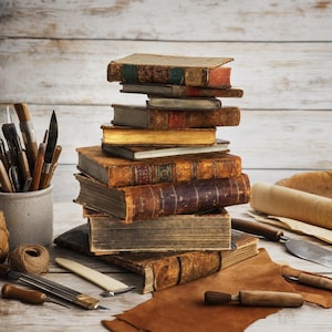 Può includere: Una pila verticale di libri antichi rilegati in pelle con bordi usurati e dorsi visibili, accompagnata da vari strumenti di legatoria tra cui punteruoli in metallo, una stecca d'osso, un rocchetto di spago e un pezzo di cuoio marrone su legno.