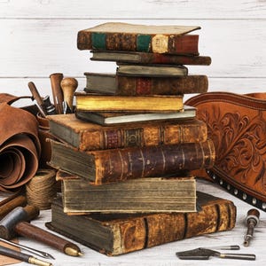 Pode incluir: Uma pilha vertical de livros antigos e desgastados com capas de couro envelhecido e bordas douradas. Ao redor dos livros estão várias ferramentas de couro, incluindo um martelo de madeira, sovelas de metal, um carretel de fio e couro lavrado.