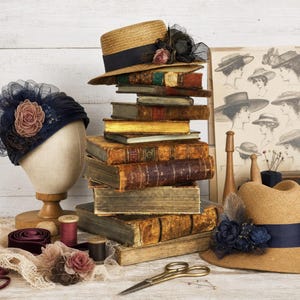 May include: A stack of antique books sits beside a navy blue fascinator on a mannequin head, a straw boater hat with a dark ribbon, a second straw hat with blue floral accents, vintage sewing spools, lace trim, and metal scissors.