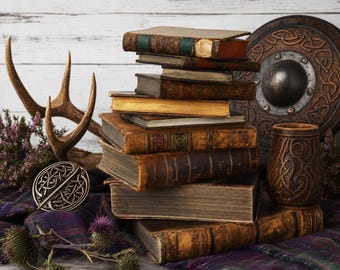 May include: A stack of weathered, leather-bound antique books sits beside a pair of deer antlers, a circular metal shield with Celtic knotwork, a carved wooden mug, a silver-toned circular brooch, and a sprig of purple thistle on a plaid fabric.