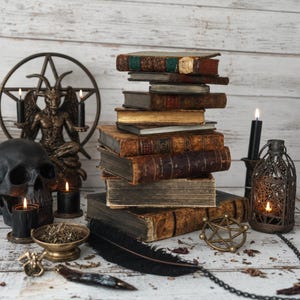 Könnte beinhalten: Ein Stapel verwitterter antiker Bücher steht in der Mitte, umgeben von einem schwarzen Totenkopf, einer bronzenen Baphomet-Statue mit Pentagramm, schwarzen Kerzen, einer Metalllaterne, einer Messingschale mit getrockneten Kräutern und einer schwarzen Feder.