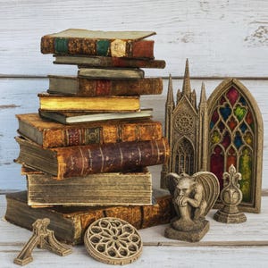May include: A stack of weathered, leather-bound antique books sits beside a stone-colored gargoyle figurine, a miniature gothic cathedral facade, a stained-glass window frame, a circular stone medallion, and a small architectural arch piece.