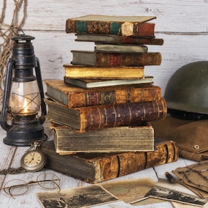 May include: A stack of weathered antique books with worn leather covers, a black metal oil lantern with a glowing flame, a silver pocket watch, round wire-rimmed spectacles, a military helmet, and several vintage black and white photographs.