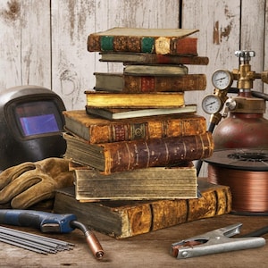 May include: A stack of antique, weathered hardcover books sits beside a black welding helmet, a metal gas cylinder with two pressure gauges, a spool of copper wire, a blue welding torch handle, metal rods, and a silver metal grounding clamp.
