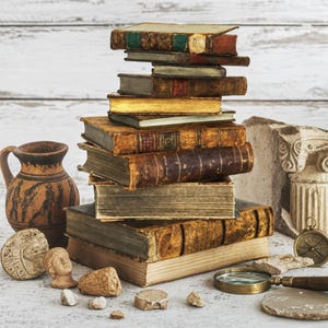 May include: A stack of antique leather-bound books with weathered pages, accompanied by a terracotta pitcher featuring black figure illustrations, a small stone bust, a brass compass, a magnifying glass, and fragments of stone architectural columns.