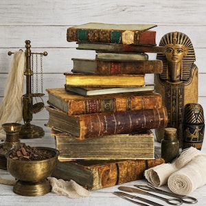 May include: A stack of antique leather-bound books sits beside a golden Egyptian sarcophagus figurine, a brass balance scale, a metal mortar filled with dark herbs, a small glass bottle, medical gauze rolls, and two metal surgical instruments.