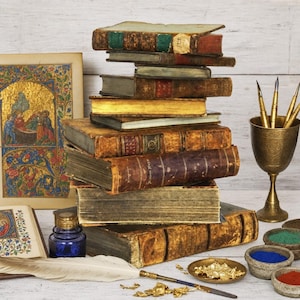 May include: A stack of antique leather-bound books with gold-edged pages, a blue glass inkwell, a white quill pen, a brass chalice holding three metal nib pens, a small dish of gold leaf, and pigment bowls next to an illuminated manuscript page.