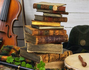 May include: A stack of weathered, antique hardback books with worn leather spines, positioned beside a wooden violin, a dark green tweed hat, a black flute, a small drum with a wooden mallet, and green clover leaves on a plaid fabric.