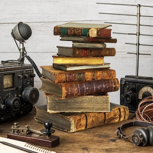 Può includere: Una pila di libri antichi con copertina rigida, dorsi in pelle consumati e pagine ingiallite, posizionata tra un trasmettitore radio vintage con microfono, un tasto telegrafico, un'antenna in metallo e un paio di cuffie classiche.