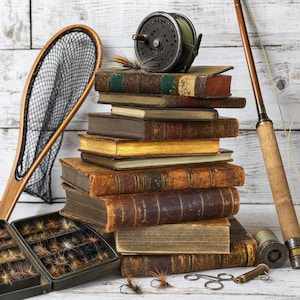 May include: A stack of weathered, leather-bound antique books topped with a dark metal fly fishing reel. Beside the books are a wooden landing net, a fly fishing rod with a cork handle, a metal fly box filled with lures, and small metal tools.