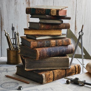 May include: A tall stack of weathered, antique hardcover books with worn leather spines and aged pages. Surrounding the books are brass drafting tools, including a compass, a divider, a metal container, and a wooden pencil on a technical drawing.