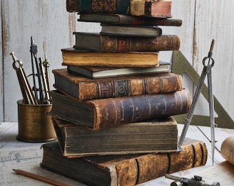Peut inclure: Une haute pile de livres anciens à couverture rigide, aux dos en cuir usé et aux pages jaunies. Autour des livres se trouvent des outils de dessin en laiton, dont un compas, une pointe sèche, un récipient en métal et un crayon en bois.