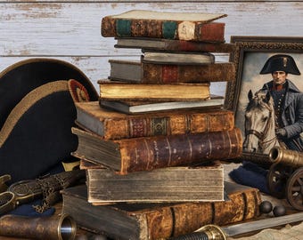 May include: A stack of weathered, antique leather-bound books sits beside a black bicorne hat, a framed portrait of a military officer on horseback, a brass telescope, a metal sword hilt, and a small model cannon on a wooden surface.