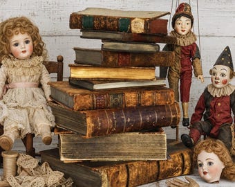 May include: A stack of antique leather-bound books surrounded by two dolls, a marionette, a doll head, a wooden hand, a paintbrush, lace trim, and metal scissors. The dolls feature detailed facial expressions and period-style clothing.