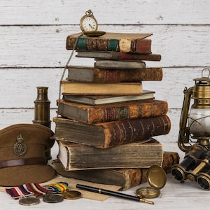 Op de afbeelding: Een stapel antieke boeken met een gouden zakhorloge, vergezeld door een bruine militaire pet, een koperen telescoop, een metalen lantaarn, een verrekijker, een zwarte vulpen en diverse ronde medailles met kleurrijke linten op wit hout.