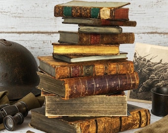 May include: A stack of antique, weathered books with worn leather spines and gold-gilded edges. Beside the books are a dark metal military helmet, vintage binoculars, two brass bullet casings, and a black-and-white photograph of soldiers.