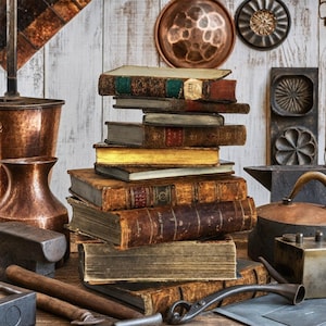 May include: A vertical stack of antique, leather-bound books with weathered spines and worn edges, surrounded by various vintage copper kitchenware, metal molds, and heavy iron tools on a rustic wooden surface with a white-washed wood background.