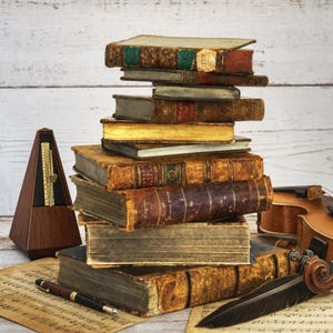 May include: A stack of antique, leather-bound books with weathered spines and gold-gilded edges, accompanied by a wooden pyramid-shaped metronome, a fountain pen, a violin, and a black feather quill resting on sheets of vintage musical notation.