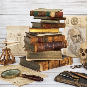 May include: A stack of antique leather-bound books sits alongside a wooden model of a flying machine, a magnifying glass, a human skull, and sketches of the Vitruvian Man and a bearded portrait, all arranged on a light-colored wooden surface.