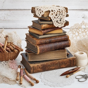 May include: A stack of seven weathered, brown hardcover books topped with a cream-coloured crocheted doily. Beside the stack are a small woven basket filled with wooden bobbins, several loose bobbins, a ball of white yarn, and metal scissors.