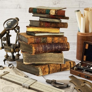 May include: A stack of antique leather-bound books sits beside a brass transit level, a wooden drafting tool case, a slide rule, and a cylindrical container holding rolled paper documents. The items are arranged on a light-colored wooden surface.