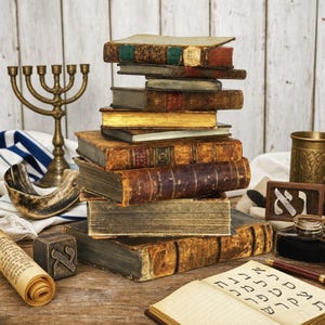Op de afbeelding: Een stapel antieke, verweerde boeken met versleten leren kaften en goudkleurige paginaranden. Naast de stapel staan een koperen menora, een sjofar, een rol met Hebreeuwse tekst, een houten blok met de letter Aleph en een open notitieboek met Hebreeuws.