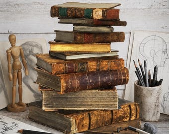 May include: A stack of antique, leather-bound books with weathered spines and gold-gilded edges. Beside the books are a wooden artist mannequin, a ceramic pencil holder filled with graphite drawing pencils, and loose charcoal sticks.