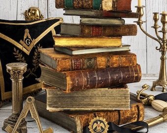 May include: A stack of antique leather-bound books sits beside a black velvet masonic apron with gold embroidery, a brass square and compass tool, a small column, a gold eagle emblem on a black ribbon, and a brass candelabra on a white surface.