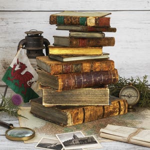Könnte beinhalten: Ein Stapel verwitterter, antiker Bücher mit abgenutzten Ledereinbänden und goldfarbenen Buchschnitten. Daneben befinden sich eine walisische Flagge, eine Metalllaterne, ein Messingkompass, eine Lupe, alte Schwarz-Weiß-Fotos und gerolltes Papier.