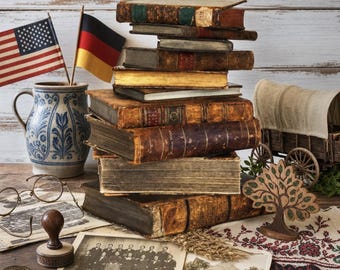 May include: A stack of antique leather-bound books, a blue and white ceramic pitcher, two small flags of the USA and Germany, wire-rimmed spectacles, a wooden stamp, a metal coin, a wooden tree cutout, and vintage black and white photographs.