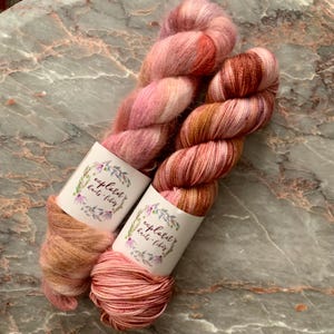 May include: Two skeins of hand-dyed yarn in shades of pink, peach, and brown. Each skein is wrapped with a white label featuring the text "explored knits + fibs" surrounded by a floral design. The yarn is displayed on a marble surface.