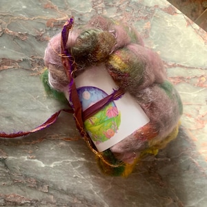 May include: A bundle of colorful wool roving in shades of pink, green, and yellow, wrapped with a purple ribbon and a card featuring a whimsical illustration of a landscape with a moon and flowers. The wool is soft and fluffy.
