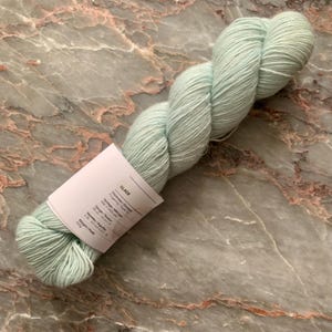 May include: A skein of Glace yarn in a soft, pale green color. The yarn is tightly wound and has a smooth texture. A white label is attached, with text that includes the word "Glace". The yarn is 100g.