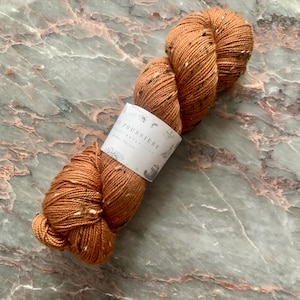 May include: A skein of orange-brown yarn with flecks of white and dark brown. The yarn is wrapped with a white label that has the text "E Poussiere" and botanical illustrations. The yarn is likely for knitting or crocheting.