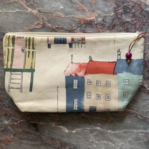 Puede incluir: Un estuche rectangular color crema con cremallera y una colorida ilustración de edificios. Los edificios tienen tejados y paredes rojos, azules y verdes. Un tirador de cuero marrón con una cuenta morada está unido a la cremallera.