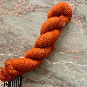 May include: A skein of orange yarn, tightly twisted, lies on a marble surface. The yarn is a vibrant, solid color, with a slight sheen. The texture of the yarn is visible, suggesting a soft, natural fiber. The overall presentation is clean and simple.