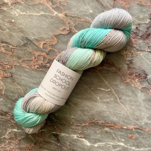 May include: A skein of Fashion School Dropout yarn in shades of teal, gray, and cream. The yarn is hand-dyed and has a speckled appearance. The label reads "Fashion School Dropout yarn co."