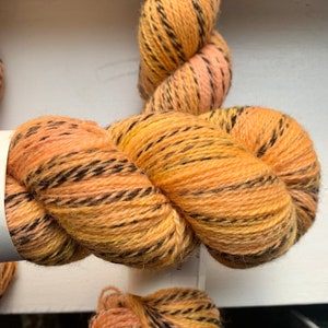May include: A skein of yarn in a peach, yellow, and black colorway. The yarn is a blend of wool and silk and has a soft, fuzzy texture.