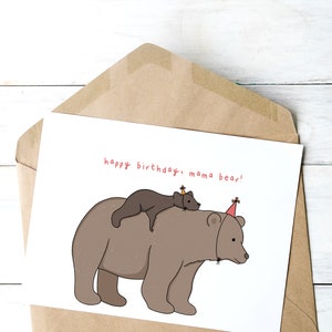 Pu&ograve; includere: Biglietto di compleanno in una busta marrone. La carta presenta un'illustrazione a cartoni animati di un grande orso marrone con un orso pi&ugrave; piccolo sulla schiena, entrambi con cappelli da festa. Il testo "happy birthday, mama bear!" &egrave; scritto in rosso.