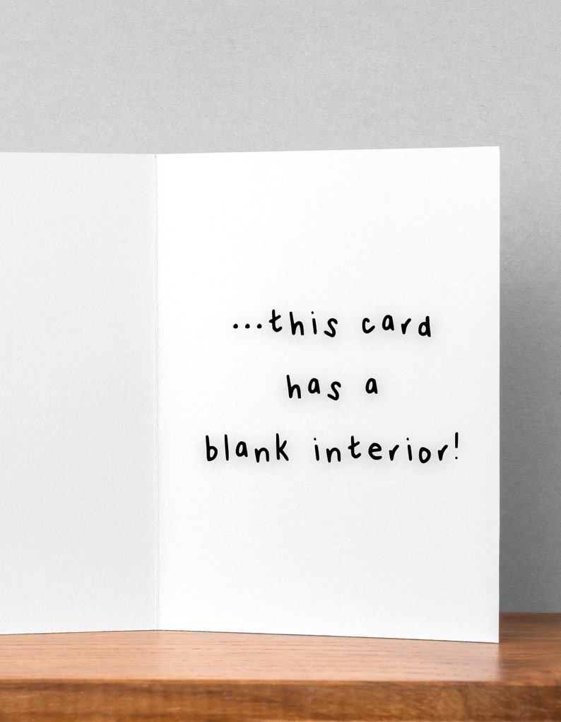 Pu&ograve; includere: Biglietto di auguri bianco aperto che rivela un interno vuoto. Il testo sul lato destro recita: "...this card has a blank interior!". Il biglietto &egrave; su una superficie di legno con uno sfondo grigio.