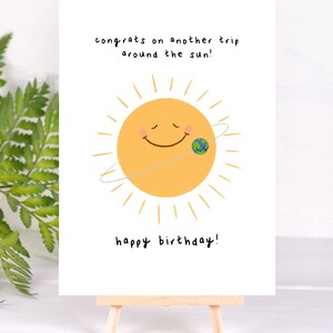 K&ouml;nnte beinhalten: Geburtstagskarte auf einer kleinen Holzstaffelei. Die Karte zeigt eine l&auml;chelnde gelbe Sonne mit einer kleinen Erdengrafik und dem Text "congrats on another trip around the sun!" und "happy birthday!"