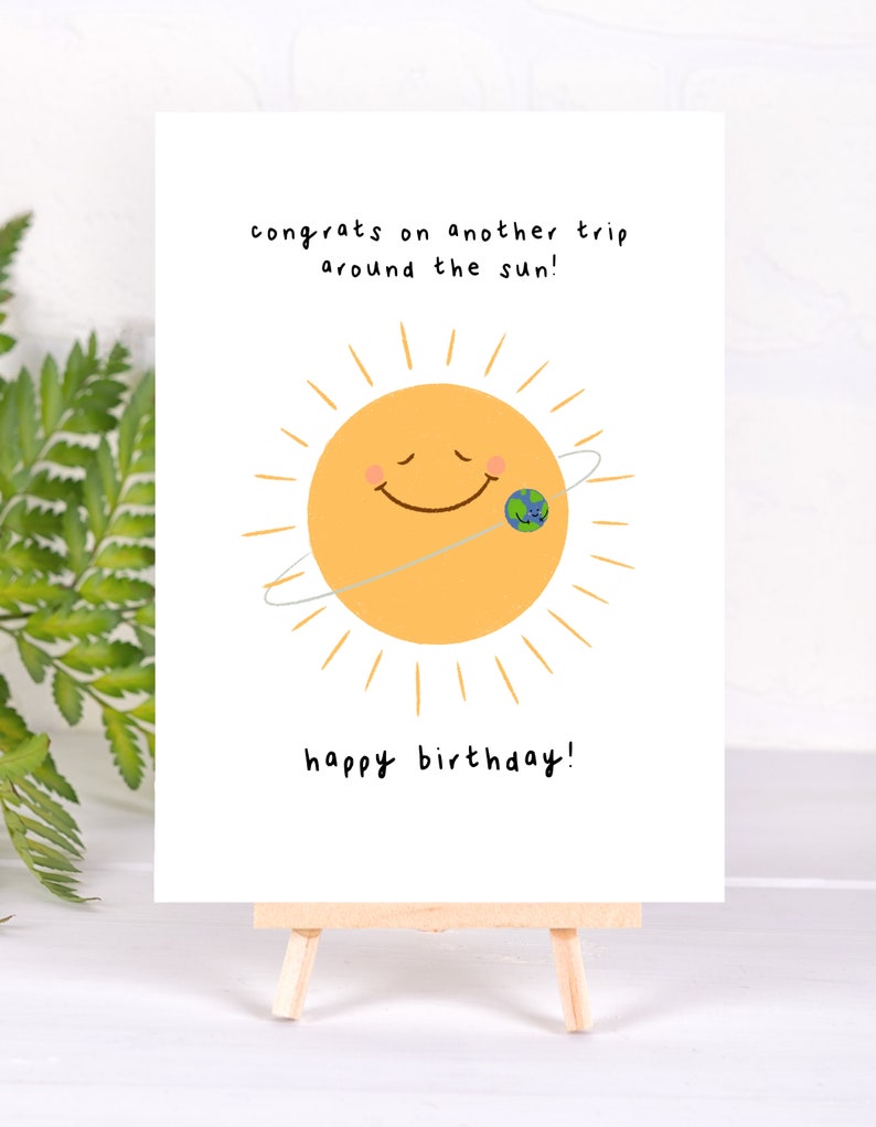 K&ouml;nnte beinhalten: Geburtstagskarte auf einer kleinen Holzstaffelei. Die Karte zeigt eine l&auml;chelnde gelbe Sonne mit einer kleinen Erdengrafik und dem Text "congrats on another trip around the sun!" und "happy birthday!"