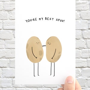 May include: A white greeting card with the text "YOU'RE MY BEST SPUD!" features two cartoon potatoes with arms and legs. The potatoes are light brown with dark brown spots and are drawn in a simple, whimsical style.