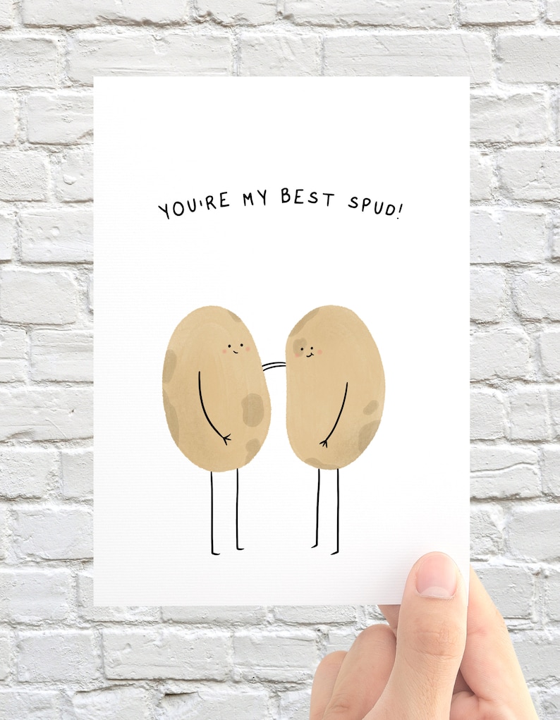 May include: A white greeting card with the text "YOU'RE MY BEST SPUD!" features two cartoon potatoes with arms and legs. The potatoes are light brown with dark brown spots and are drawn in a simple, whimsical style.